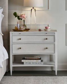 a nightstand with two books on it next to a lamp and flowers in a vase