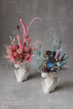 two white vases with flowers and feathers in them on a gray surface, one is holding the other's head