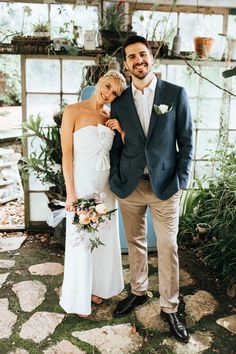 a bride and groom pose for a photo in front of a greenhouse with potted plants