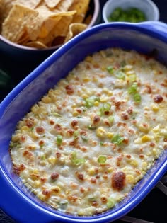 a blue casserole dish filled with corn, cheese and green onions next to chips