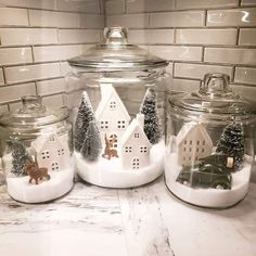 three glass jars with houses and trees in them on a countertop next to a tile wall