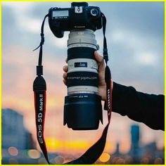 a person holding up a camera in front of a cityscape at dusk with the sun setting behind them