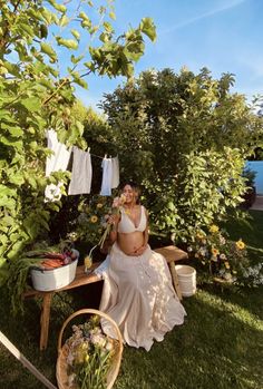 a pregnant woman sitting on a bench in the grass next to some flowers and clothes