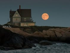 the full moon is setting over a house on top of a cliff near the ocean