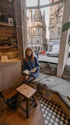 a woman sitting on a bench in front of a window reading a book while looking out at the street