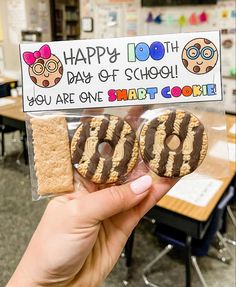 a person holding up two cookies in front of a sign that says happy 100th day of school you are one smart cookie