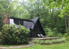 a black cabin in the woods surrounded by trees and bushes with lounge chairs on the lawn