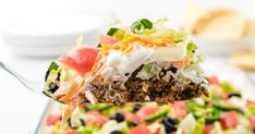 a close up of a spoon full of food with salad in the background and chips on the side