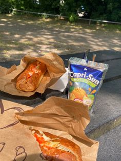 two hotdogs in brown paper bags sitting on a bench next to a bag of chips