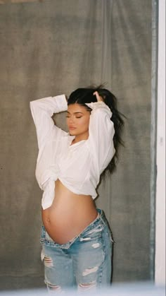 a pregnant woman in ripped jeans and a white shirt poses for the camera with her hands behind her head