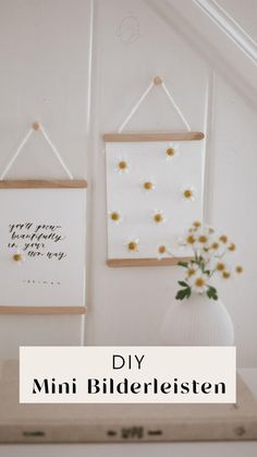 two framed pictures hang on the wall above a vase with daisies in it and a white vase with flowers