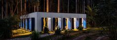 a small white building with blue shutters in front of some trees and bushes at night