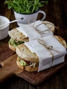 two sandwiches wrapped in white paper and tied with twine on a cutting board next to a bowl of greens