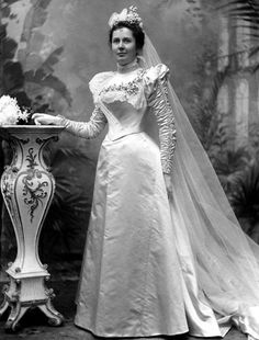 an old black and white photo of a woman in a wedding dress standing next to a vase