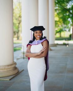 a woman wearing a graduation cap and gown standing in front of columns with her arms crossed