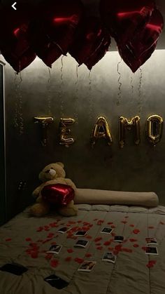 a teddy bear sitting on top of a bed next to red heart shaped balloons hanging from the ceiling