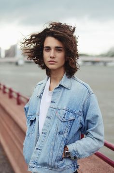 a man with long hair standing in front of a fence and looking at the camera