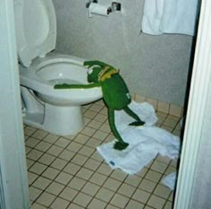 a green frog sitting on top of a toilet in a bathroom next to a roll of paper