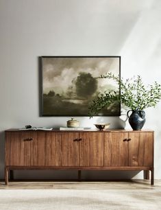 a painting hanging on the wall next to a wooden cabinet with two vases and a plant