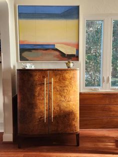 a wooden cabinet sitting in front of a window next to a painting on the wall