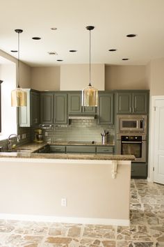 a kitchen with green cabinets and marble counter tops, an island in the middle is surrounded by hanging lights