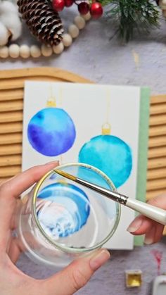 someone is holding a paintbrush and painting christmas ornaments on a card with watercolors