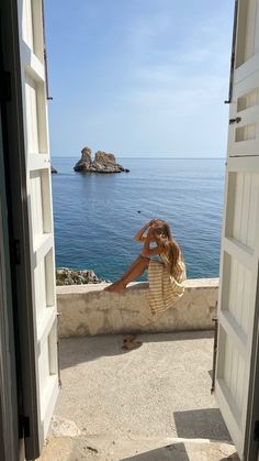 a woman sitting on top of a stone wall next to the ocean with an open door