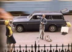 a man standing next to a woman in front of a black car with a camera