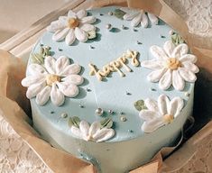 a blue cake decorated with white flowers and the word happy written on it is in a cardboard box