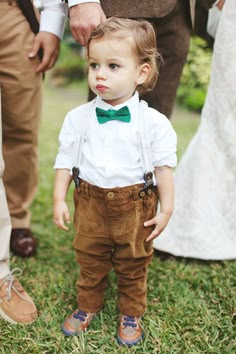 a little boy wearing a bow tie and suspenders standing in the grass with other people