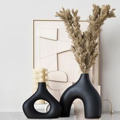 two black vases sitting next to each other on a white counter top with dried plants in them