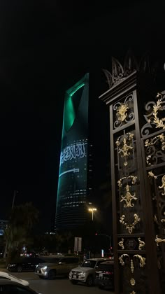 a tall building lit up at night with cars parked in the parking lot below it