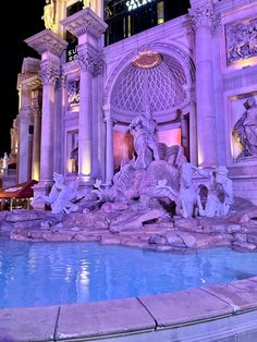 a fountain in front of a large building with statues on it's sides and lit up at night