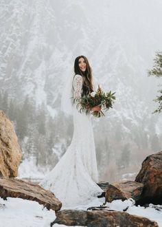 a woman in a wedding dress holding a bouquet standing on snow covered rocks with mountains in the background