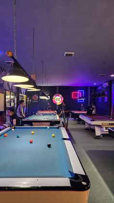 several people playing pool in a large room with neon lights on the ceiling and tables