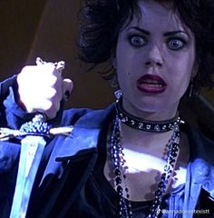 a woman with dark hair and blue eyes holds a knife in her hand while wearing an elaborate necklace