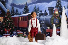 a young boy standing in front of a christmas scene with a suitcase and teddy bear