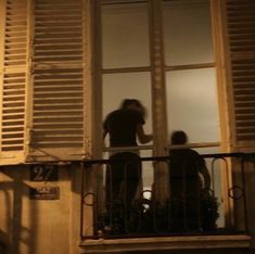 two people standing on a balcony looking out the window at night with shutters open