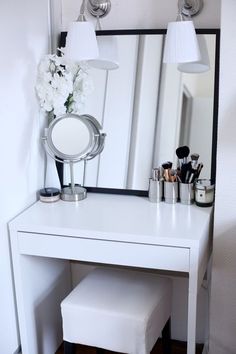 a white vanity table with a mirror and some makeup brushes on it, next to a flower vase