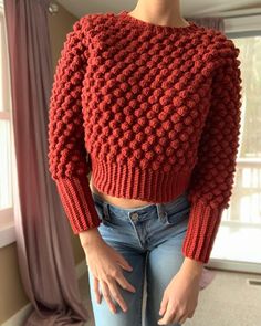 a woman is standing in front of a window wearing a red knitted sweater and jeans