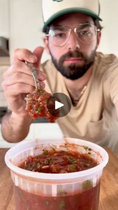 a man holding a spoon full of food in front of a container filled with sauce