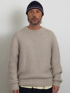 a man standing in front of a white wall wearing a gray sweater and black hat