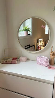 a white dresser topped with a round mirror next to a pink purse and potted plant