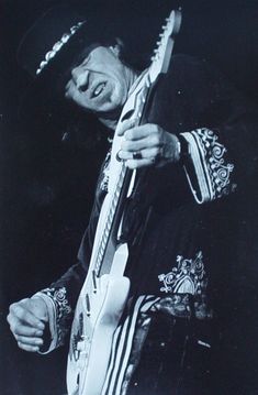 a black and white photo of a man holding a guitar in his right hand while wearing a hat