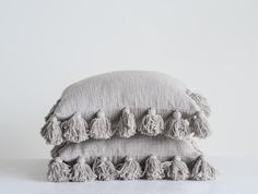 two grey pillows with tassels on them sitting side by side in front of a white wall