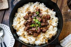 a skillet filled with rice and meat on top of a wooden table next to utensils