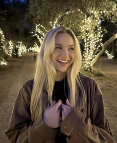 a woman standing in front of some trees with lights on the ground and smiling at the camera