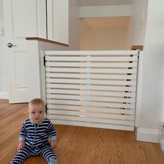 a baby sitting on the floor in front of a white gate with horizontal slats