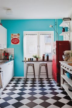 a black and white checkered floor in a small kitchen with blue walls, an old fashioned refrigerator and two stools