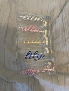 three clear glasses with different colored writing on them sitting on a white bed sheet in front of a pillow